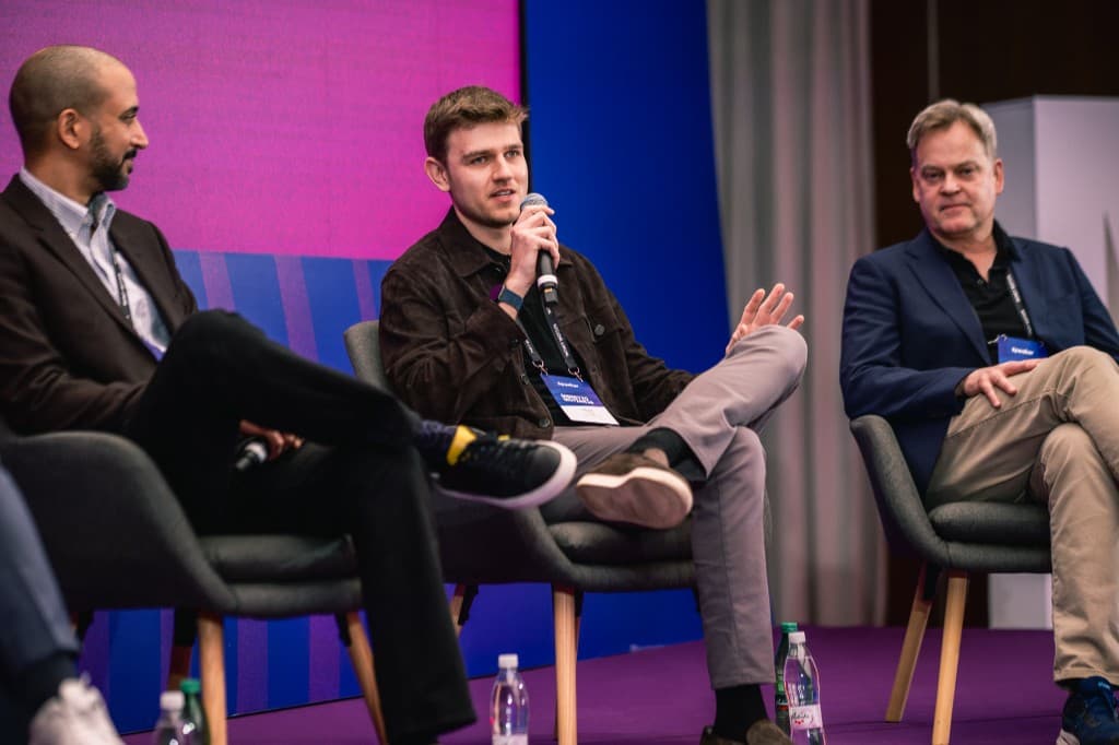 Three people seated on stage in discussion, colorful gradient backdrop and speaker lanyards